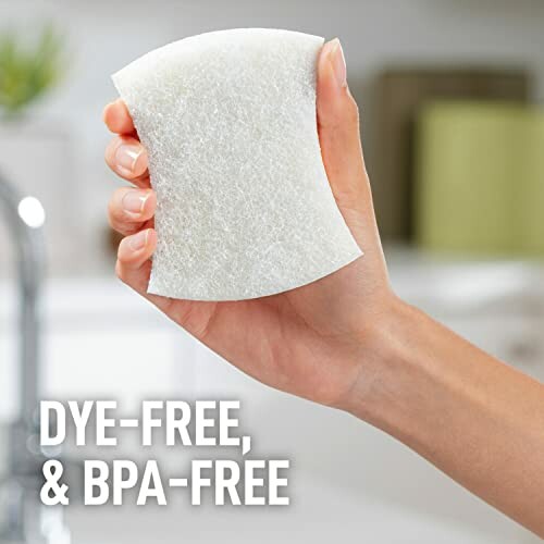 Hand holding a dye-free and BPA-free sponge in a kitchen setting.