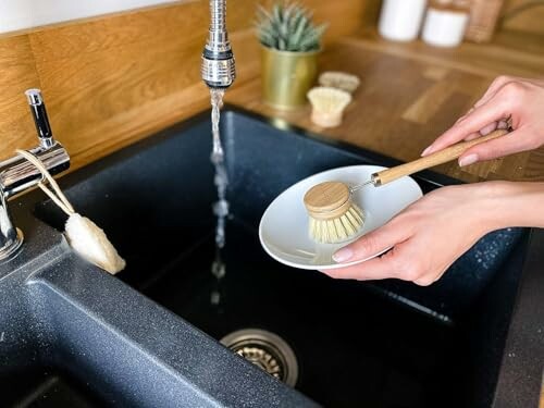 Person using a dish brush to clean a plate under running water in a kitchen sink.