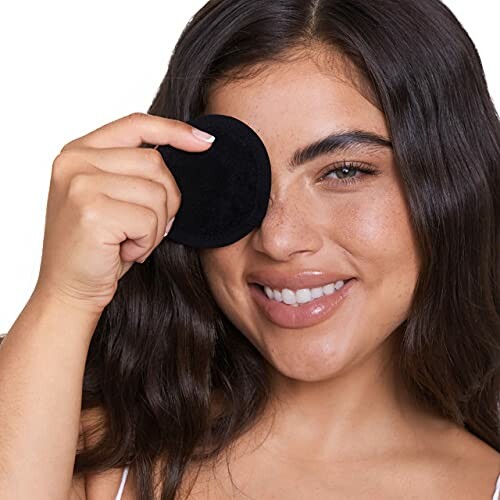 Woman smiling while holding a black makeup remover pad to her eye.