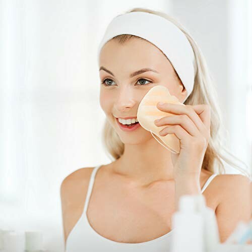 Smiling woman using a facial sponge in front of a mirror.